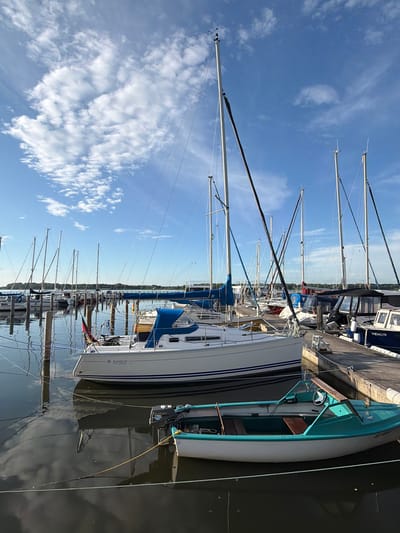 This media file shows a marina with numerous sailboats docked in Ribnitz-Damgarten, Germany. The scene is captured on a clear day with scattered white clouds in a bright blue sky, suggesting it is likely daytime, possibly late morning or afternoon. The water is calm, reflecting the boats and the sky, indicating a peaceful atmosphere.
The primary subjects are the sailboats, with their masts tall and prominent against the sky. One sailboat in the foreground has a white hull with blue stripes and a dark-colored jib. Another teal-colored boat is partially visible. Several other sailboats are docked alongside a concrete pier, lined up neatly.
There are no people visible in the frame. The activity is static, with the boats at rest in the harbor. Notable details include the rigging of the sailboats, the concrete dock, and the reflections in the water. One of the boats has the text "VENUS" and "JEANNEAU" visible on its hull, along with some smaller print that is not clearly legible. The overall impression is one of a serene maritime setting.