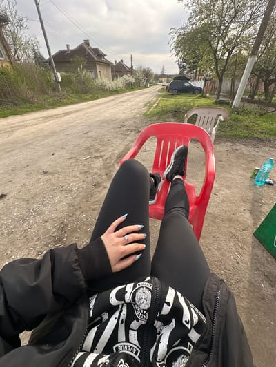 The image depicts a person seated on a red plastic chair outdoors in what appears to be a rural or semi-rural area. The person is wearing black leggings, black sneakers, and a black jacket with a white and black patterned inner layer. The person's left hand is visible with blue-and-white nail art and a ring on the ring finger. The ground is dirt and gravel. In the background, there is a dirt road, houses, and vegetation, including trees. A white plastic chair is to the left. A dark car is parked in the background. Based on the location clues provided, this scene is set in Galiche, Bulgaria. A plastic water bottle and other litter are in the top left of the frame. The perspective is from above, suggesting the photo was taken from the seated person's point of view.