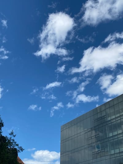 A modern glass-faced building in Sainte-Anne-de-Bellevue, Canada, is captured from a low angle, showcasing its reflective facade against a bright blue sky dotted with fluffy white clouds. The building's geometric lines and glass panels dominate the right side of the frame, reflecting the surrounding environment and sky. On the left, a portion of a building with a reddish-tiled roof and some greenery, including trees and branches, are visible. The scene is devoid of any people.
