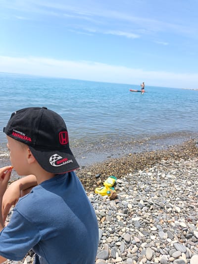 The image presents a sunlit pebble beach scene in Krinitsa, Russia, along the Black Sea coast. In the immediate foreground, positioned to the lower left and center, a young male child is observed from his left profile and back, seated on the multi-colored pebbles. He wears a dark blue short-sleeved t-shirt and a black baseball cap. The cap features "POWERED by HONDA" embroidered in red on the front-left panel, a red Honda "H" logo on the side, and "HONDA RACING" with a checkered flag design on the brim. His left hand is raised to his mouth, holding a small, light-colored food item, possibly a nut or cracker.
The beach itself comprises a dense expanse of smooth, rounded pebbles ranging in shades of gray, white, and brown, extending from the foreground into the midground. Near the child's right, partially obscured by pebbles, rests a yellow plastic toy, possibly a sandal or small boat, alongside a piece of driftwood.
The calm, blue-green sea extends across the midground to the horizon under a clear, light blue sky scattered with faint, wispy white clouds. In the distant midground, a lone individual stands on a red paddleboard, actively paddling, positioned roughly in the center of the visible water. The lighting is bright and natural, indicating a sunny day, with subtle shadows cast by the boy and pebbles.