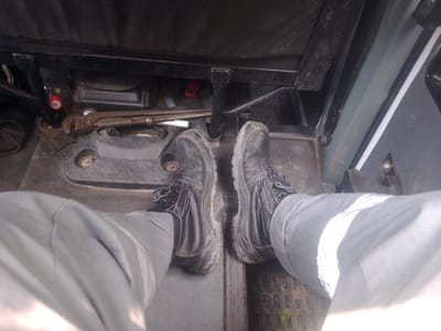 The image shows a first-person perspective of a person's feet and lower legs inside what appears to be a vehicle cabin, possibly a tractor or construction equipment. The person is wearing dark-colored work boots with laces and light gray work pants. The floor of the cabin is made of textured black material. In the background, there are various metal components and what looks like a wrench or tool. The environment is dimly lit. There is no indication of the city of Yessentuki, Russia in the image.