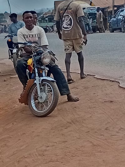 The image depicts a roadside scene in Ayangba, Nigeria. In the foreground, a light-skinned male, approximately 20-30 years old, is seated on a motorcycle, facing slightly left of center and smiling towards the camera. He wears a light-colored t-shirt with dark text, possibly reading "WAKEEL USMAN," and dark trousers. His left hand rests on the motorcycle's handlebar. The motorcycle is light blue/silver with a chrome headlight and spoked wheels, parked on a reddish-brown dirt shoulder.
To the right and slightly behind the seated male, a dark-skinned male, also approximately 20-40 years old, walks away from the camera, facing towards the right. He wears a tan t-shirt with a prominent graphic on the back featuring a stylized face and the text "HELP STAR." He also wears tan shorts and open-toed sandals. He holds a small, light-colored object in his left hand.
The background shows a paved road with multiple vehicles, including other motorcycles, cars, and a light-colored van or truck. Several other individuals are visible, some standing by the road, others seemingly in transit. Structures with reddish-brown roofs are present in the distance. The sky appears overcast. The immediate ground in the foreground is unpaved, consisting of reddish-brown dirt, indicating a roadside area adjacent to a paved street.