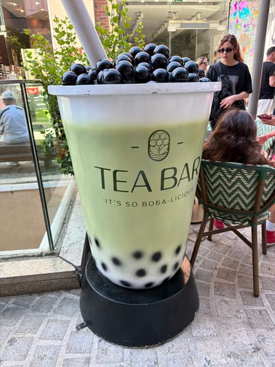 The image depicts a large, green, oversized cup of bubble tea, a popular beverage. The cup is prominently placed outdoors, on a cobblestone-like surface, likely a sidewalk or plaza. The branding on the cup reads "TEA BAR" with the tagline "IT'S SO BOBA-LICIOUS". Black boba pearls are visible both at the top of the drink and at the bottom. A large white straw is also inserted into the cup.
In the background, several individuals are visible. To the right, a woman with long, wavy brown hair, wearing sunglasses and a black t-shirt with a gray graphic, is looking towards the left. She has a dark bag slung across her body. Behind her, another person, possibly male, is partially visible wearing a black t-shirt. To the left of the giant bubble tea cup, a person in a white t-shirt and green stripe jacket is seated on a bench, facing away from the camera. Storefronts with glass windows are also visible in the background. The overall setting appears to be an urban environment, consistent with a commercial or pedestrian area in the city of Ashdod, Israel. The lighting suggests it is daytime.