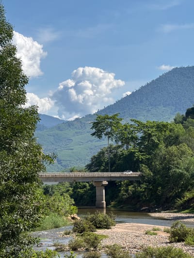 An outdoor, eye-level shot captures a bridge spanning a river in Nam Khanh Vinh, Vietnam. The bridge is made of concrete and supported by substantial pillars, with a car visible driving across it. Lush green trees flank the river, with a dense forest and rolling, verdant mountains forming the background. The sky above is a clear blue, dotted with fluffy white clouds. The river flows over a rocky bed, with some vegetation growing along its banks. The overall scene suggests a tranquil, natural landscape enhanced by human infrastructure.