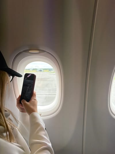 This image provides an intimate view from an airplane window seat, likely during daytime. The primary subject is a person, seen from the chest down, holding a dark-cased smartphone up to the oval-shaped aircraft window. Their right hand grips the phone, which appears to be dark or powered off, reflecting the bright exterior light. Long, light-colored hair of the person cascades on the right side of the frame, and a leg or foot wearing a black shoe is visible near the bottom left of the window.

The interior of the airplane cabin is characterized by smooth, light grey walls and ceiling, featuring a subtle horizontal groove or light strip running across the upper section. Through the window, a bright outdoor scene is visible, showing elements of green foliage and light-colored structures, suggesting an airport environment or landscape. The illumination from outside indicates it is daytime. The activity captured is a common one during travel: a passenger interacting with their phone while looking out the window, possibly taking a photo or video. Given the location context, this scene likely takes place at or near Tillé, France. No discernible text is visible in the image.