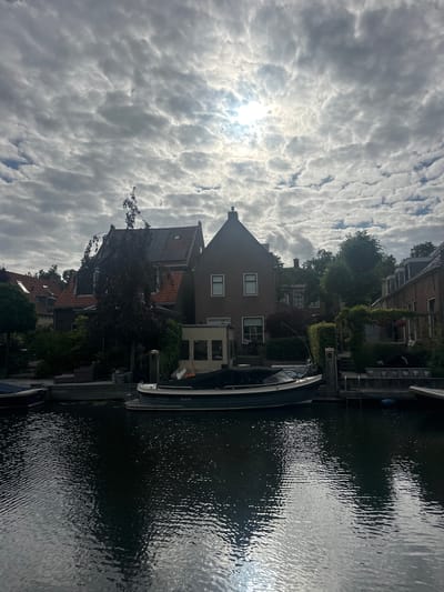 The image shows a canal in Katwijk, Netherlands, lined with houses and trees. Several boats are docked on the canal, and the water reflects the overcast sky. The sun is partially visible through the clouds, casting a bright spot on the water's surface. The scene appears to be a quiet, residential area.