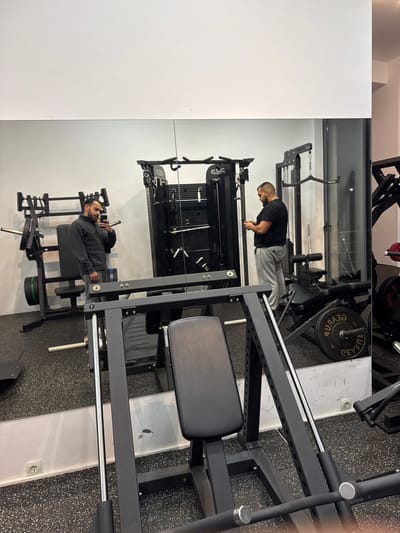 This image captures an interior view of a fitness facility, identified as a gym, located in Plataniás, Greece. The floor is uniformly covered with black, speckled rubber gym flooring. The walls are white, with a prominent mirror covering the lower half, reflecting the interior space.
In the immediate foreground, a black leg press machine, featuring a padded seat and silver support rods, is partially visible. To the left within the mirror's reflection, a male individual with dark hair and a beard is seen wearing a dark grey quarter-zip jacket and grey sweatpants. He holds a smartphone in his right hand, angled towards the mirror, indicating he is either taking a photograph or recording video of himself. Behind him, reflected gym equipment, including a machine with green and black weight plates, is visible.
To the right, standing directly in the gym space in front of the mirrored wall, another male individual with dark, short hair and a beard is present. He is wearing a black short-sleeved t-shirt and light grey sweatpants, holding a smartphone with both hands and looking down at its screen. Behind him and to his left, a black multi-functional cable machine, branded "SFS" in its reflection, is visible. Other black gym equipment, including weight racks and machines with reflected red weight plates, are distributed throughout the background. A white electrical outlet is visible on the wall in the reflection near the floor.