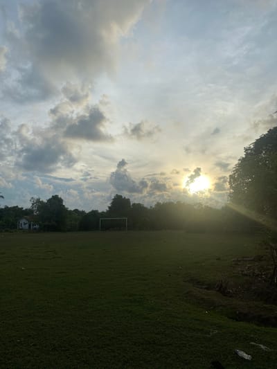 The image depicts an outdoor scene in Keudeublang, Indonesia. The sky is filled with dramatic clouds, with the sun breaking through, casting rays of light and creating a bright, luminous effect. The time of day appears to be late afternoon or early evening, given the angle of the sun and the quality of light. The weather seems to be partly cloudy, with a mix of bright sunshine and darker cloud formations.
In the foreground and midground, there is a large expanse of green grass, likely a field or park. Trees line the background, creating a natural border. A soccer goal is visible on the field, suggesting it might be a sports ground. A small, white building is also present near the treeline. The overall mood is serene and natural, with a hint of the approaching end of the day. There are some scattered pieces of litter, including a plastic bottle and some string, on the grass, which are minor details in the otherwise picturesque landscape. No people are visible in the image. There is no visible text.