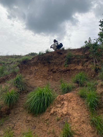 The image displays a person navigating a steep, earthy slope under an overcast sky. The individual, seen from a low angle, appears to be a male wearing a light-colored (possibly white or light grey) long-sleeved top and dark trousers, with a dark backpack or container strapped to their back. They are positioned on the brown soil and rock face, which is interspersed with clumps of bright green grass and some sparse leafy plants. The person's body language suggests they are carefully moving along or climbing the incline. Above, the sky is filled with grey, heavy clouds, indicating an overcast day. The terrain is rugged and natural, characteristic of an undeveloped, rural, or hilly area. No text is visible. The location context provided is Amaigbo, Nigeria.