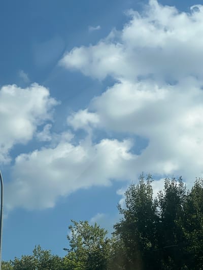 The image shows a bright blue sky with scattered white cumulus clouds. The sun is not visible, but the lighting suggests it is daytime. There are some trees with green foliage visible at the bottom of the frame, appearing to be part of a park or a residential area. A vertical gray pole, possibly a street light or utility pole, is partially visible on the left side of the image. The scene is likely outdoors in an urban or suburban setting. The location is specified as Chirshkasy (Sirmaposinskogo S/P), Russia.