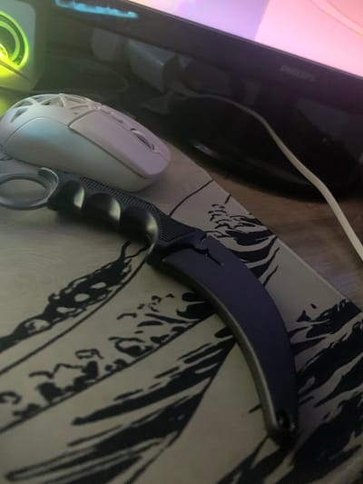 A karambit knife with a dark purple blade and a black handle rests on a patterned mousepad. To the left of the knife is a white gaming mouse with a honeycomb design. In the background, a dark monitor with a PHILIPS logo is visible. The scene appears to be a desk setup. The image does not provide any information about the location in Glodeni, Moldova.