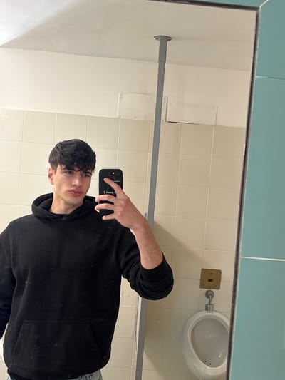 A young male is captured in a mirror selfie taken inside what appears to be a public restroom. He is positioned slightly to the right of the frame, looking directly into the smartphone camera held in his right hand. He has dark, wavy hair and is wearing a black hoodie and light-colored jeans, partially visible at the bottom right. His expression is neutral.

The setting is a tiled room, likely a men's restroom, given the visible white porcelain urinal with a brass-colored flush plate mounted on the wall to its left. The lower part of the wall is covered with light beige or cream-colored square tiles, while the upper section, visible in the mirror's reflection, is a solid turquoise or teal color. A metallic horizontal bar, possibly a support rail or part of a partition, runs across the middle of the reflection. Various rectangular panels are visible on the tiled wall, suggesting access points or fixtures. The lighting is artificial, typical of an indoor public space.

On the back of the black smartphone case, the text "GOOD (:" is visible in reverse due to the mirror reflection. The image is oriented vertically (portrait mode), but the original capture was likely landscape and then rotated, showing the scene on its side. The general location is Camerino, Italy.