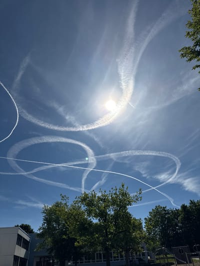 The image depicts a bright, sunny day with a clear blue sky featuring numerous contrails from aircraft. The contrails form intricate, looping patterns, with some appearing to intersect. The sun is visible, slightly obscured by a hazy cloud formation and a lens flare, suggesting it is mid-morning or early afternoon. In the foreground and along the right side of the frame, lush green trees with dense foliage are visible, some of which are casting shadows. Behind the trees, parts of a modern building complex with windows are discernible. The scene appears to be set in an outdoor urban or suburban environment, possibly a park or a residential area, given the presence of trees and buildings. There is no visible text in the image. The overall impression is one of a dynamic sky over a serene, leafy setting.