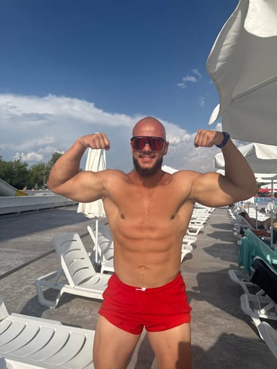 A bald man with a beard and sunglasses is posing by flexing his biceps in a resort setting. He is wearing red swim trunks and is shirtless. The background shows rows of white lounge chairs, white umbrellas, trees, and a blue sky with clouds, suggesting a sunny day at an outdoor recreational facility. The man is the primary subject, and his muscular physique is prominent. The scene appears to be a typical resort or water park environment. There is no specific indication that this location is in Ivanyane, Bulgaria, beyond the prompt's directive.
