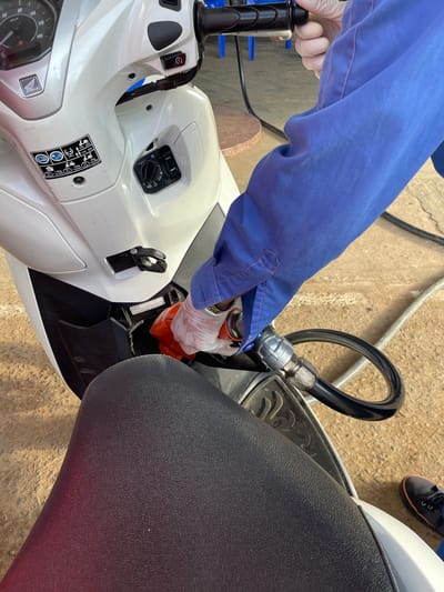 A detailed overhead shot captures the process of refueling a white Honda scooter at a gas station in Cam Ly - Da Lat, Vietnam. The right arm of an individual, clad in a blue long-sleeved shirt with orange stitching and a clear disposable glove, is positioned on the right side of the frame, holding an orange and silver fuel pump nozzle. The nozzle is inserted into the scooter's front-mounted fuel port. The scooter's white bodywork is prominent, featuring a silver Honda logo and a partially visible dashboard with a speedometer indicating up to 140 km/h. A black informational sticker with white icons, including weight limits (e.g., "175 328 lbs"), is affixed to the left side of the front console. Below the dashboard, a keyless ignition system with a rotary switch and a "FUEL" button is visible. A black cargo hook is present above the fuel port. The textured black seat of the scooter occupies the lower half of the image, from the rider's perspective. The ground is a light brown concrete surface with visible lines. A dark blue shoe with an orange sole is partially visible at the bottom right, next to the black fuel hose connecting to the nozzle.