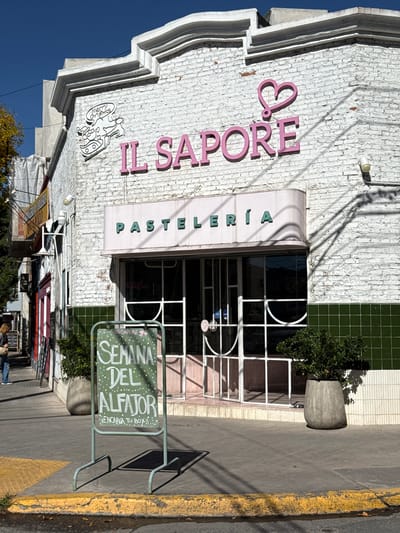 This exterior shot reveals a "Il Sapore" patisserie in Salta, Argentina. The business name, rendered in pink cursive, is visible on a white brick building front. A storefront awning reads "PASTELERIA" in green, above glass doors and windows that display the patisserie's interior. A sidewalk sign advertises "SEMANA DEL ALFAJOR," promoting alfajores, a traditional Argentine treat. Plants in concrete pots flank the entrance. A yellow curb defines the street edge and parked vehicles can be seen. People are also seen walking along the street. The sky is clear and blue.