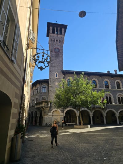 The image depicts a sunlit square in Bellinzona, Switzerland. On the right side of the square, a large, stone building with arched windows and a covered walkway is visible. A mature tree with green foliage stands in front of the building. To the left, a narrow street leads away from the square, flanked by buildings with balconies. A tower, likely part of a historical structure, rises above the buildings, featuring a clock face with Roman numerals. A decorative metal sign depicting a deer hangs from a pole extending from the building on the left.
In the foreground, a woman wearing a black and white patterned jumpsuit walks away from the camera, carrying a black bag. She is on a cobblestone pavement. In the background, another person is partially visible near the arches of the building on the right, and a bicycle is parked in the distance. The sky is clear blue. The overall scene suggests a historical town center during daytime.