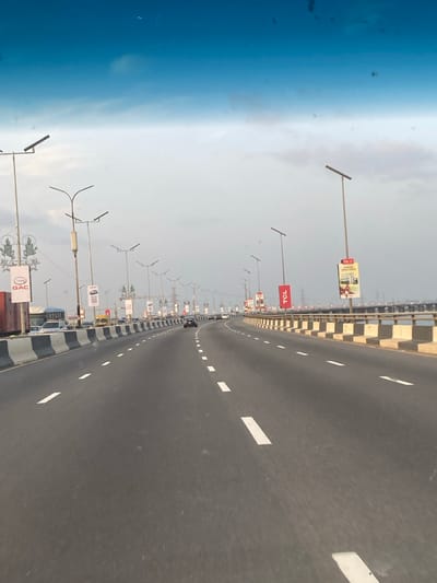 A highway view reveals a long stretch of asphalt, marked with white lane dividers. A black and white striped barrier runs alongside the road, accompanied by numerous light poles and billboards, some displaying the "TCL" logo. The sky transitions from a clear blue to a hazy gray, suggesting either the early morning or late afternoon hours. Cars appear in the distance, indicating traffic flow on the bridge.
