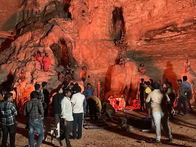 The image depicts a scene set within a natural cave environment, illuminated by a prominent orange or red light source, likely artificial lighting for a film shoot. The cave walls are textured and rugged, with visible geological formations. Several individuals are present, appearing to be part of a film crew or actors.
In the background, on elevated rock ledges, are figures dressed in what appear to be traditional Indian attire, possibly depicting religious or historical characters. One figure on the left is in a red garment, and another further back on a ledge also wears red. To the right, several figures are seated on ledges, also in similar attire. A ladder is visible leaning against the cave wall in the middle-right section, suggesting access to higher areas.
In the foreground and mid-ground, a group of people are gathered. Some are dressed in contemporary clothing, such as jeans and t-shirts, suggesting they are crew members. One individual in a grey and black plaid shirt is standing with his back to the camera, looking towards the cave interior. Another person in a white shirt and dark pants is also visible. A film set apparatus, likely a dolly or camera track, is set up on the ground in the mid-ground.
The overall impression is a controlled environment within a cave, possibly for a film or television production, with actors in costume placed strategically on the natural rock formations and a crew managing the logistics in the foreground. The specific location in Gollagutta, India, is noted, implying a geographical context for this scene.