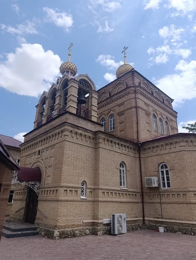 The image shows an exterior view of a brick church with a golden dome and cross, set against a blue sky with scattered clouds. The church features arched windows, a bell tower with multiple bells, and decorative architectural details. In the foreground, a paved courtyard is visible, along with an air conditioning unit and a broom leaning against the wall. The overall scene depicts a religious building in a clean, well-maintained environment. The church is identified as the Church of the Resurrection of Jesus Christ, located in Ashgabat, Turkmenistan.