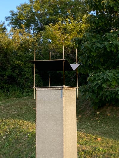 A weathered, galvanized metal structure with a dark, flat roof supported by thin, metallic poles stands outdoors. A small, white, triangular piece of material is affixed to one of the poles near the roof. The structure is positioned in front of a dense backdrop of green trees and foliage, with a clear blue sky visible above. The ground in front of the structure is covered with green grass, with some scattered small white flowers. The overall scene appears to be in a natural, possibly rural or park-like setting. The structure does not have any discernible markings or context to indicate its specific purpose or location within Arandjelovac Municipality, Serbia. There are no people or animals present in the image.