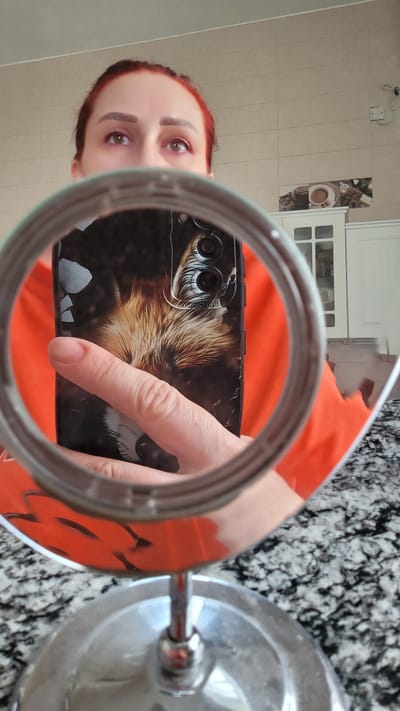 A woman with red hair is reflected in a round magnifying mirror. She is looking into the mirror, and her eyes are visible. She is holding a smartphone with a fox-themed case, which is also reflected in the mirror. The background of the reflection shows a kitchen with light-colored tiles and white cabinets. The woman is wearing an orange shirt. The image appears to be taken indoors, likely in a residential setting within the city of Ras Al Khaimah, United Arab Emirates.