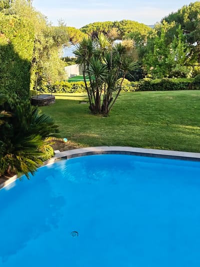 The image depicts a sunlit, manicured garden with a vibrant blue swimming pool in the foreground. A well-maintained lawn dominates the midground, bordered by a hedge and lush green trees. In the background, a body of water, likely a lake or the sea, is visible, framed by more trees and a clear sky. A small, multi-colored ball rests on the grass near the pool's edge. A stone well with a metal apparatus sits in the left midground, surrounded by greenery. The scene evokes a sense of tranquility and leisure. This location is situated in Ramatuelle, France, known for its picturesque landscapes and proximity to the Mediterranean coast.