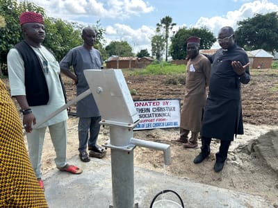 In Afogo, Nigeria, five men stand around a recently installed water well. The man on the far left wears a maroon cap and a dark vest over a light-colored garment. Next to him, a man in a grey uniform looks towards the well. The third man from the left, in a brown robe, stands with his arm around the man in the black robe on the far right. The man on the far right, possibly a clergyman, gestures with his hands while wearing glasses and a black clerical garment. Behind them, a sign indicates the well was "DONATED BY: [UNREADABLE] CHRI[UNREADABLE] SOCIATION(NCA) SUPPORT FROM:- [UNREADABLE] OF LIFE CHURCH LAGOS NIG." The background shows rural buildings with thatched roofs and trees under a cloudy sky. The well has water flowing from its spout into a bucket.