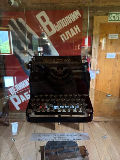 A vintage black typewriter is displayed on a glass shelf in a museum. The typewriter has a metal frame, black keys with Cyrillic lettering, and a carriage return lever. Behind the typewriter, a red banner with white lettering and a large gray hand graphic is visible. The text on the banner appears to be in Russian. To the right of the typewriter, a wooden door with a sign reading "ЗАПАСН. ВЫХОД" (Emergency Exit) is visible. The floor beneath the display is made of light-colored wood planks. A small descriptive plaque is placed in front of the typewriter, also on the glass shelf. The plaque contains Russian text. The overall environment suggests an indoor exhibition space with rustic wooden walls.