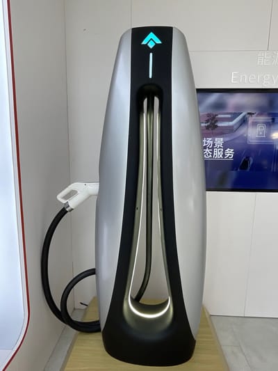 The image shows a silver and black electric vehicle charging station in a modern, white-walled space. The charging station is shaped like a teardrop with a black center containing a glowing white light and a black cable with a white connector that is plugged into the charger.  The charging station is located in the city of Hangzhou, China, with coordinates of 30.272 latitude and 120.416 longitude. There is a white cable leading to a white charging gun, which is in the foreground, disconnected from the charger. The background shows part of a white wall with a black and silver vertical panel behind the charging station.  A flat-screen monitor is mounted on the wall and displays a graphic of a parking lot with a car and an electric car charging symbol in Chinese.  The Chinese text on the screen appears to be "Energy Service," and there are several dark gray lines running vertically down the surface of the charging station.  The image is captured from a slightly oblique angle, looking straight at the front of the charging station.