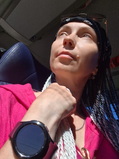 A close-up, low-angle shot shows a young woman with light skin and blue eyes, wearing a black bandana and sunglasses on her head. She has braided hair with white and gray streaks. She is wearing a bright pink top and a silver chain necklace with a gold pendant. A black smartwatch is on her left wrist. The background suggests she is in a vehicle with a dark blue seat and a dark ceiling. The lighting indicates it's daytime, with sunlight casting shadows. The image composition is centered on the woman's face and upper body, with her gaze directed upwards. The location, Красный Ключ, Russia, is not visually represented in the image.