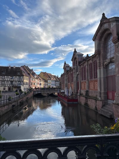 The image depicts the Lauch river in Colmar, France, a picturesque canal lined with historic buildings. On the left side, a row of half-timbered houses with colorful facades and steep roofs are visible, characteristic of the Alsatian architectural style. A stone bridge spans the canal, with people walking across it. On the right side of the canal, a large, imposing stone building with arched windows and red shutters stands prominently. Several closed white umbrellas are set up in front of this building, suggesting a cafe or restaurant. The canal itself is dark and reflective, mirroring the sky and the surrounding architecture. The sky above is partly cloudy, with patches of blue sky visible between dramatic white clouds. A black metal railing runs along the bottom of the foreground, partially obscuring the view of the canal and the buildings. The overall scene conveys a sense of charming European urban landscape bathed in natural light.