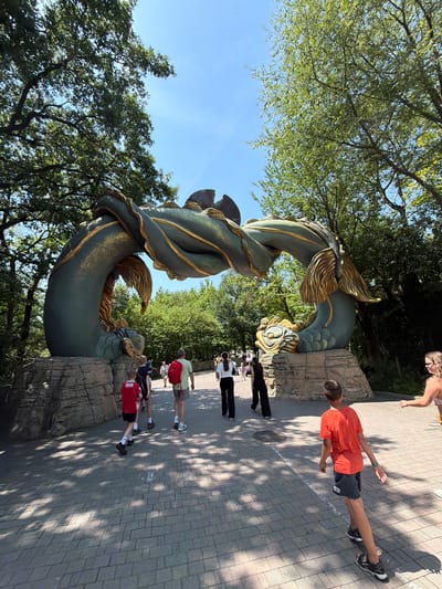 An outdoor, daytime, wide-angle shot captures an ornate archway shaped like a serpentine dragon, adorned with gold accents, set against a backdrop of lush green trees and a clear blue sky. The dragon arch is supported by faux rock structures on either side. A paved pathway, made of grey rectangular stones, leads towards and under the arch. Sunlight filters through the trees, casting dappled shadows on the pathway.

Several individuals are walking on the pathway. In the foreground, a boy wearing an orange t-shirt and dark shorts walks away from the camera. To his left, a boy in a red shirt and black shorts walks away. Behind him, a man with a red backpack and grey shorts walks away. Further ahead and to the left, a group of people, including a woman in black pants and a white shirt, are walking towards the arch. On the right side of the pathway, a woman with blonde hair wearing a light-colored top and yellow shorts is walking away from the camera.

The location is identified as Gardaland, an amusement park situated in Castelnuovo del Garda, Italy.