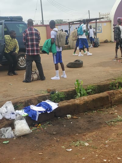 The image depicts an outdoor scene, likely near a school or institution, in what appears to be the city of Bahit, Nigeria. Several young individuals, dressed in what seem to be school uniforms, are present, some carrying backpacks. One individual is wearing a checkered shirt and appears to be interacting with someone inside a van. Another person is holding a green jacket. In the foreground, a variety of items are scattered on the ground, including plastic bags, crumpled paper, clothing items (some appear to be school uniforms in blue and white), and food waste. A discarded tire is also visible on the road. The environment appears to be unpaved, with visible soil and some vegetation growing in a trench or drainage area. The overall impression is one of a somewhat disorganized gathering area, potentially a bus stop or entrance to a school.