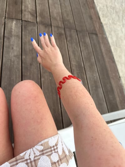 A close-up, high-angle shot shows a person's arm and hand, adorned with bright blue fingernail polish and a red, wavy bracelet, resting on a wooden deck. The person's skin is fair with freckles and some minor redness. Part of their legs and a patterned towel are also visible. The background consists of the textured wooden planks of the deck and a light-colored surface, possibly sand or pavement, to the right. The scene is set outdoors, likely in Yuzhnyye Koryaki, Russia, suggesting a warm-weather location.