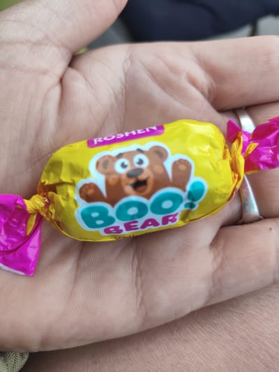 The image shows a hand holding a yellow candy wrapper with pink ends. The wrapper has a cartoon bear image and the words "Boo! Bear" printed on it, along with "Roshen". A silver ring is visible on one of the fingers. The background is blurred, showing a dark navy fabric. The image appears to have been taken in Saksahan, Ukraine, as indicated by the provided location.