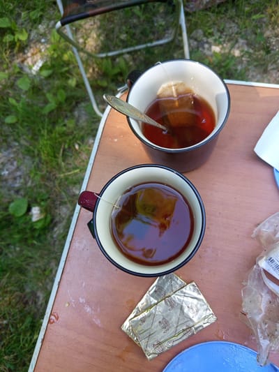 Two mugs of tea are sitting on a wooden table. One mug is in the foreground and has a tea bag in it. The other mug is behind it and also has a tea bag in it, along with a spoon. There are two packets of something shiny and gold, likely wrappers for food items, on the table. A blue plate is also visible. The table appears to be outdoors, as grass and foliage are visible in the background, as well as a portion of a folding chair. The overall scene suggests a casual outdoor setting, possibly a picnic or camping trip, with no specific indication of the city of Giaginskaya, Russia.