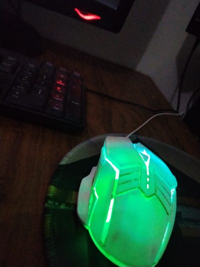 A gaming mouse with bright green LED lighting sits on a mousepad, angled towards the viewer. To the left, a portion of a black keyboard is visible, with some keys also illuminated in red. The scene appears to be on a wooden desk. The overall lighting is dim, with the primary light sources being the LEDs on the mouse and keyboard, and a red light emanating from the top left of the frame, possibly from a monitor. The location is identified as Yangibazar, Uzbekistan.