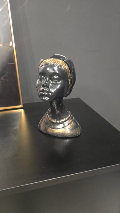 A black, bust sculpture of a woman with gold accents sits on a black table. The sculpture depicts a woman's head and neck, with her eyes closed and a serene expression. She wears a gold headscarf and a gold necklace. To the left of the sculpture is a portion of a framed artwork with a gold frame, its contents are not visible. The background is a plain, dark grey wall. The image is taken from a slightly elevated angle, focusing on the sculpture. The environment appears to be indoors, possibly a room in a house or gallery. The location is not specified in Trikomo, Northern Cyprus / Cyprus.