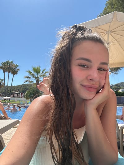 A young woman is lying on a lounge chair outdoors, likely at a resort or pool area in Andratx, Spain. She is looking directly at the camera, with her right hand resting on her cheek and chin. She has long, brown hair that falls around her face and shoulders. She is wearing what appears to be a white garment. The background reveals a bright blue sky, palm trees, and a swimming pool with people in it, suggesting a sunny day. The lighting and shadows indicate it is likely daytime, possibly late morning or afternoon. The overall mood appears relaxed and happy.