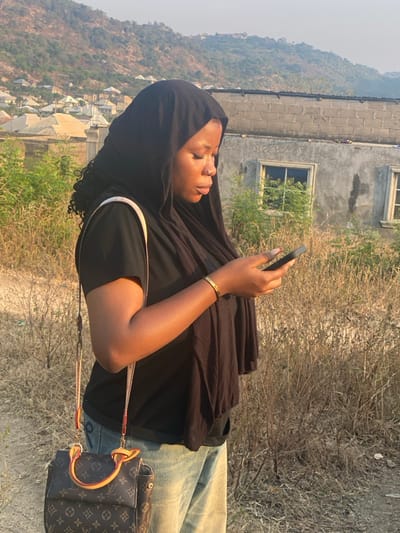 A woman with dark skin and a black hijab holds a smartphone, looking at it. She wears a black top, light-wash jeans, and a gold bracelet. A Louis Vuitton handbag hangs off her shoulder. The background includes a building, dry vegetation, and a distant hillside with residential structures. The picture is rotated 90 degrees, with the woman appearing on the left side.