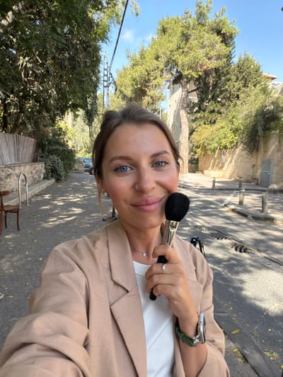A single adult female is depicted in a selfie-style image on Rehov Dubnov 18, West Jerusalem, Israel, under bright daylight. The female, approximately 25-35 years old, has light skin, blue eyes, and light brown hair pulled back. She wears a beige blazer over a white ribbed top, a silver necklace with a heart pendant, a silver ring on her left ring finger, and an Apple Watch with a dark green band on her left wrist. She holds a large makeup brush with black bristles and a metallic handle against her right cheek with her right hand, smiling subtly at the camera. The background shows an asphalt street with dappled sunlight and shadows from numerous tall evergreen trees that line both sides. On the left, a light brown wooden chair is positioned near a low stone wall topped with wooden slatted fencing. On the right, a multi-story building constructed of light-colored Jerusalem stone, partially covered by green climbing vegetation, is visible. Three dark cylindrical bollards are spaced along the right curb, and a metal grate is present in the street pavement. A dark-colored vehicle is partially visible in the distant background on the left, and a camera tripod is partially visible on the ground to the right of the woman.