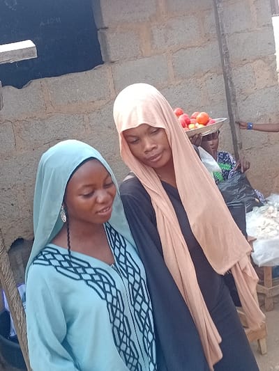 Two women in headscarves and traditional attire are positioned closely together. The woman at the top wears a peach-colored headscarf and a black outfit. She holds a tray of red fruits. The other woman, located below, has on a pale blue headscarf and a matching top with decorative trim. The background is slightly blurred, revealing part of a stone structure and other indistinct elements.