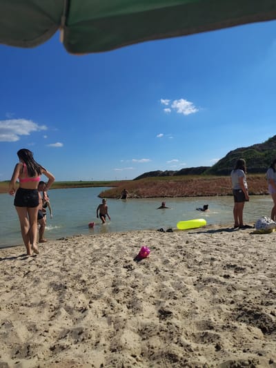 A group of people are enjoying a sunny day at a beach, likely a lake or river, in Shumeikiv, Ukraine. The scene is set on a sandy shore with clear blue skies and a few scattered clouds. The sun appears to be high in the sky, suggesting it is midday.
Several individuals are in the water, either swimming or wading. One person is using a bright yellow inflatable tube. On the shore, a young woman in a pink bikini top and black shorts is posing with her hand on her head, her long dark hair wet. Beside her, a man in black shorts is partially submerged in the water. In the background, other figures can be seen enjoying the water and the beach.
The landscape features a hilly, vegetated area bordering the water. The sand appears textured and dry in some areas, with some shadows cast by the sun. A pink inflatable toy is visible on the sand. The overall mood seems relaxed and recreational, with people taking advantage of the warm weather for swimming and enjoying the outdoors. No visible text is present in the image.