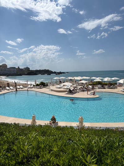 This image captures a vibrant, sunny scene at a coastal resort, identified as Biarritz, France. The foreground is dominated by a large, curving swimming pool filled with clear, blue water, its surface rippled by light. To the right of the pool, a dense border of lush green foliage runs along its edge.
Numerous people are visible enjoying the facilities. Several individuals are in the pool, some swimming, others standing in the water. On the spacious pool deck surrounding the water, more people are relaxing on white lounge chairs, many shaded by crisp white umbrellas. One person, wearing a striped top, is seated by the pool's edge, seemingly reading or engaged with a book or device. Another individual in light attire is partially visible behind the foliage near the pool.
Beyond the pool area, a sandy beach stretches towards the ocean. The calm, blue sea extends to the horizon, dotted with gentle waves near the shore. In the distance, along the coastline, various buildings and natural formations are visible under a bright blue sky filled with scattered white, wispy clouds.
The image depicts a leisurely daytime scene, suggesting pleasant weather and a relaxed atmosphere. There is no visible text.