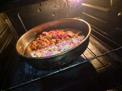 A pizza is being cooked in an oven. The pizza is in a round pan and appears to have ham and cheese as toppings. The oven rack is visible, and the interior of the oven is dark with the light from the oven illuminating the pizza. The scene is indoors, likely in a kitchen. The specific location in Kaleytsa, Bulgaria, cannot be determined from the image.