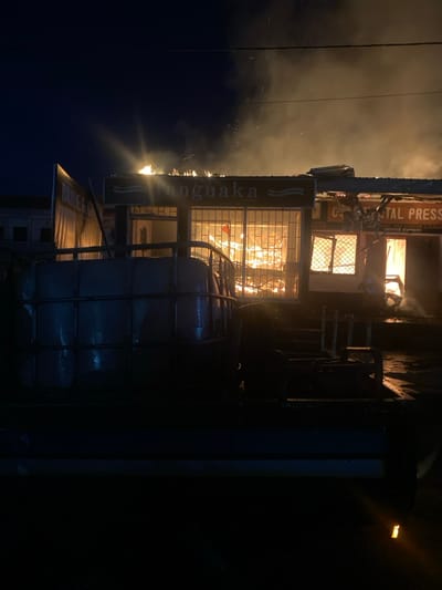 The image shows a building engulfed in flames at night. The building appears to be a commercial establishment, with a sign that reads "Ongnaka" above the windows. The building is located at the coordinates 4.011, 9.215, which is in the city of Limbe, Cameroon. The flames are bright and intense, and smoke is billowing from the roof of the building. There is a large, dark object in the foreground, possibly a truck or van, obscuring the front of the building. There is another sign visible in the background that reads "Continental Press." It is dark outside and there are no people visible in the image. The scene appears to be a fire in progress.