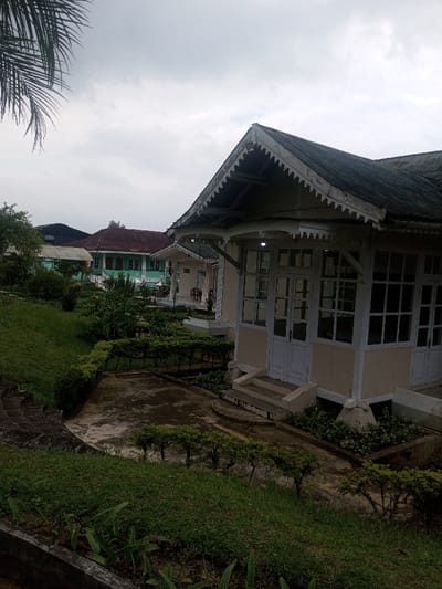 The scene is set in Nghĩa Đồng Commune, Vietnam, under an overcast sky. Dominating the right foreground is a single-story building of colonial architectural style, featuring light beige walls, white window frames, and white double doors. Its dark, gabled roof is adorned with weathered white scalloped bargeboards. The building is elevated on a foundation with concrete steps leading to its entrance. To the left, a landscaped area includes an upward-sloping grassy lawn, concrete pathways, and low, manicured hedges and small trees. In the midground, additional buildings are visible, notably one with a reddish-brown roof and a light green facade, positioned behind the foreground structure. Further back, another light-colored building can be seen. A palm frond partially obscures the top left corner of the frame. No individuals, actions, or interactions are depicted.