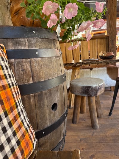 The image depicts an outdoor seating area with rustic wooden furniture and decorative elements. In the foreground, a portion of a weathered wooden barrel is visible, adorned with black metal bands. A plaid pillow, featuring orange, black, and white checks, rests against the barrel.
Behind the barrel, a small, round stool with a fur-covered seat and three wooden legs stands on a wooden floor. Further back, a wooden table with a natural wood edge is set with a small, dark brown bowl containing a golden object. A rustic wooden fence forms a backdrop behind the table.
Hanging above the table are pink petunias in bloom, cascading from a wooden planter. In the mid-ground, benches with white and brown cowhide upholstery are visible. The overall atmosphere suggests a cozy, rural, or garden setting, possibly in Shiroka Laka, Bulgaria, known for its traditional architecture and natural beauty. No people are present in the image.