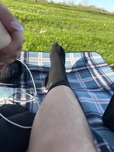 The image shows a person sitting on a blue and white plaid blanket in a grassy area, presumably outdoors. The person is wearing black socks. A white cable (likely a charging cable or headphones) is present on the blanket near the person's feet. The background includes green grass and trees. It is unknown whether the location is Beli Brod, Bulgaria.